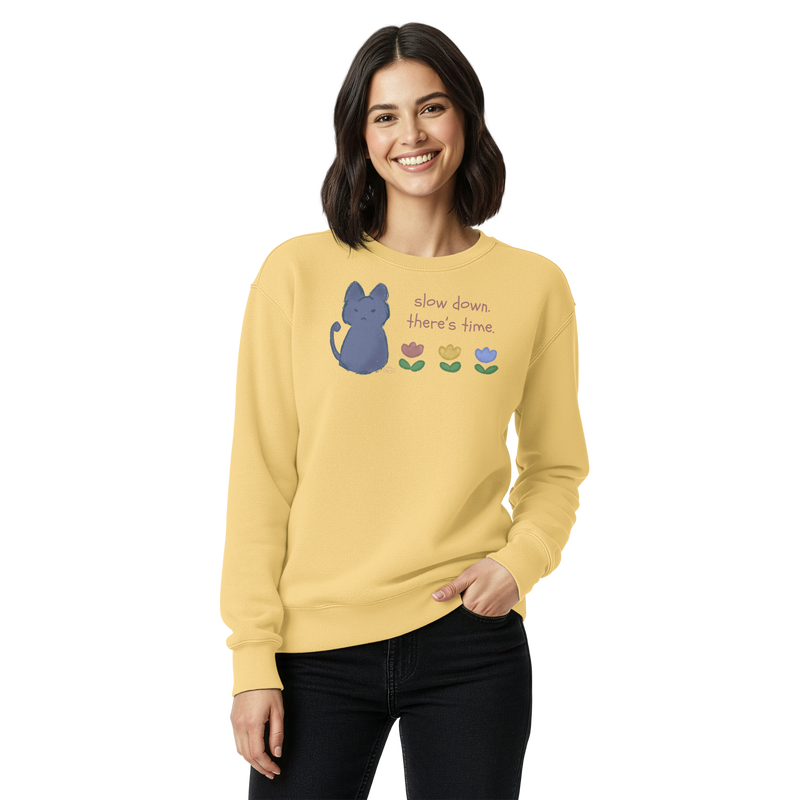 Model wearing a butter yellow garment-dyed sweatshirt with a blue cat, three flowers, and 'Slow down, there's time' lettering.