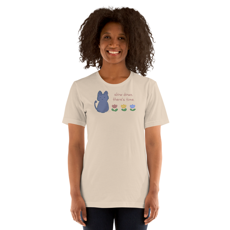Model wearing a soft cream Bella + Canvas T-shirt with a blue cat, three flowers, and 'Slow down, there's time' lettering.